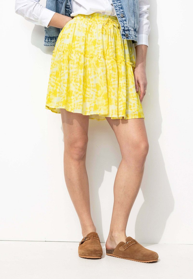 Jupe à volants jaune en tissu léger et fluide. Dotée d'une taille réglable avec cordon et d'un motif aquarelle. Associée à des chaussures décontractées en daim brun.
