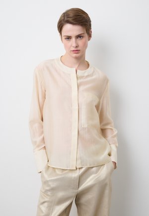 Junge Person mit kurzen Haaren trägt eine transparente cremefarbene Bluse und glänzende hellgoldene Hosen, stehend vor einem schlichten weißen Hintergrund.