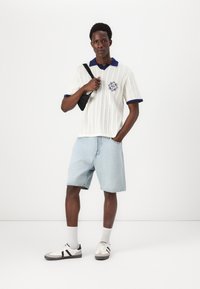 Polo de punto blanco con cuello y puños en marino; pantalones cortos de mezclilla; bolso cruzado negro; zapatillas grises con detalles negros; la modelo se encuentra de pie sobre un fondo neutro.