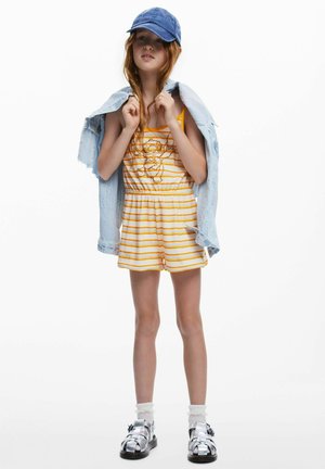 Gelb-weiß gestreifter Strampler mit einem aufgestickten Charakter, Jeansjacke lässig über die Schultern gelegt, blaue Mütze und silberne Sandalen mit weißen Socken.