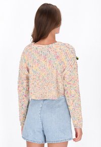 Frau, die einen mehrfarbigen, grob gestrickten, cropped Pullover und hellblaue, hochgeschnittene Jeansshorts trägt, mit dem Rücken zu einem weißen Hintergrund gewandt.