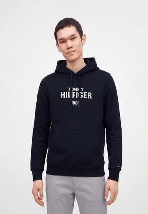 Young man wearing a black Tommy Hilfiger hoodie with white logo, standing with hands relaxed by his sides against a plain background.