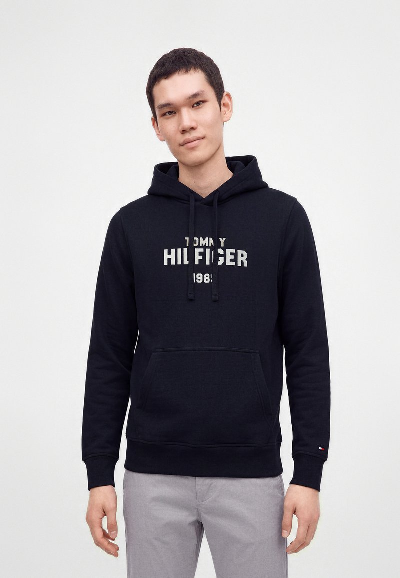 Jeune homme portant un sweat à capuche noir Tommy Hilfiger avec un logo blanc, debout les mains détendues le long du corps devant un fond uni.