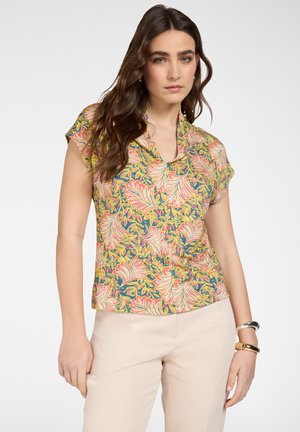 Donna con capelli castani lunghi che indossa una camicetta gialla a maniche corte con motivo floreale e pantaloni beige chiaro, con braccialetti al polso destro.