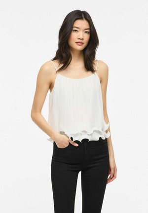 Jeune femme aux cheveux noirs mi-longs portant un haut blanc sans manches plissé et un pantalon noir, debout devant un fond uni.