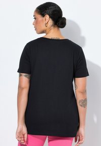 Camiseta negra de manga corta con cuello redondo, textura suave y corte holgado. Tatuajes visibles en los brazos del modelo.