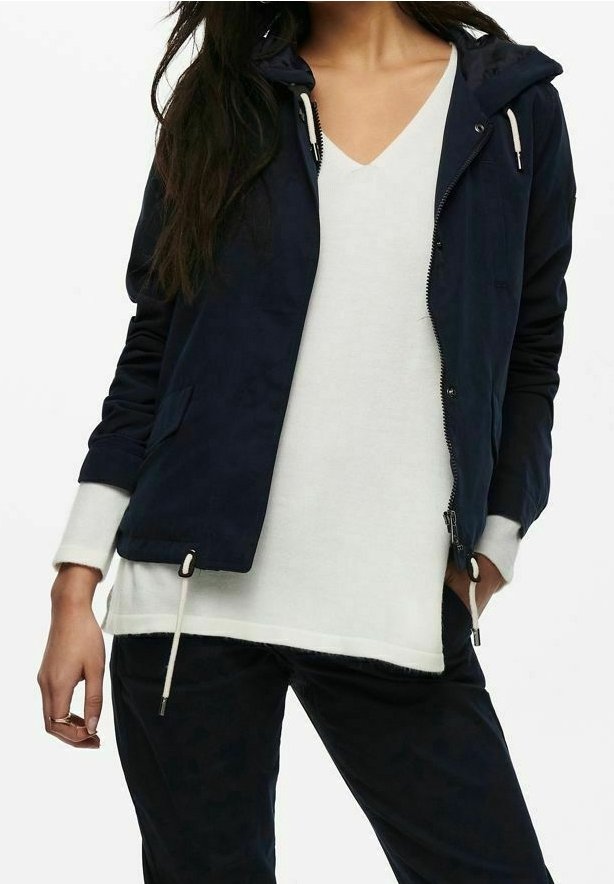 Giacca leggera blu navy con cappuccio, zip frontale, dettagli con coulisse e due tasche laterali, abbinata a una maglietta bianca con scollo a V.