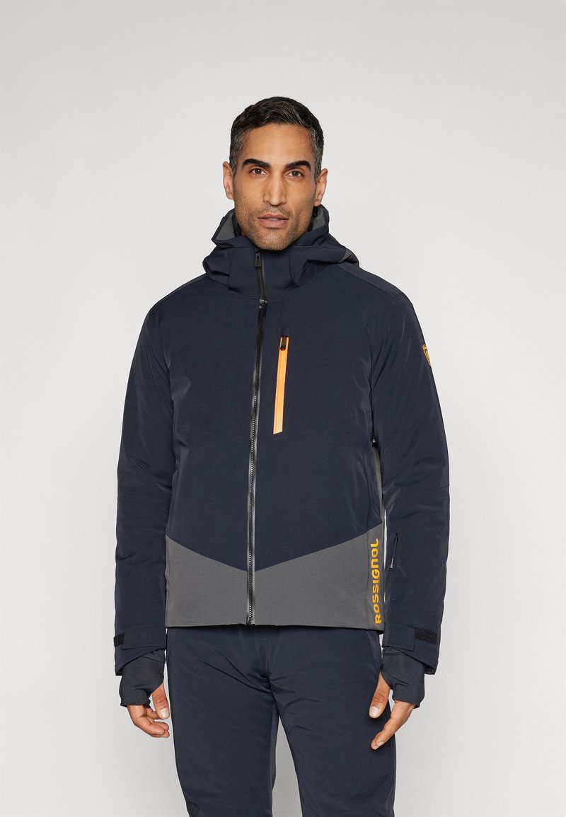 Uomo con giacca da sci navy e grigia con zip arancione e logo "Rossignol", in piedi davanti a uno sfondo grigio chiaro uniforme.