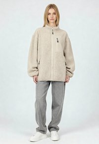 Giacca in pile beige con collo alto, cerniera frontale e tasche laterali; dotata di una tasca piatta sul petto e maniche comode. Indossata con pantaloni grigi e scarpe da ginnastica bianche.