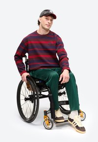 Csíkos pulóvert és zöld nadrágot viselő férfi ül egy kerekesszékben, baseball sapkát és sportcipőt visel, egyszínű háttér előtt.