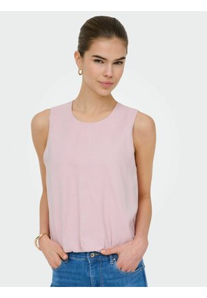 Kobieta ubrana w bladoróżowy, bezrękawnik i niebieskie dżinsy, ręce w kieszeniach, ułożone włosy oraz złote kolczyki-koła i bransoletka.