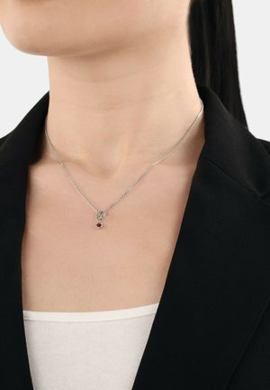 Collana in argento con un piccolo pendente circolare caratterizzato da un accento di pietra preziosa rossa, indossata su una maglietta bianca, sopra un blazer scuro.