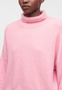 Femme portant un pull à col roulé en maille rose douce avec une coupe décontractée, visible du cou jusqu'au milieu du torse sur un fond uni.