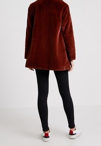 Brązowy płaszcz z sztucznego futra o luźnym kroju, z długimi rękawami i płaskim kołnierzem, zestawiony z czarnymi legginsami i białymi tenisówkami z czerwonymi akcentami.