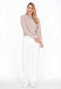Pulover din fir tricotat de culoare bej deschis, cu mâneci lungi, asortat cu pantaloni albi cu talie înaltă și croială largă, purtat cu adidași albi, stând pe un fundal simplu.