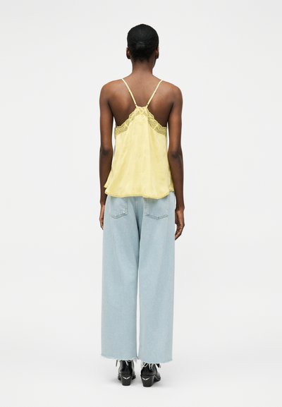 Γυναίκα με σκουρόχρωμο δέρμα και μαλλιά σε κότσο, που φοράει ένα κίτρινο τοπ με δαντέλα, ανοιχτό μπλε τζιν με φαρδιά πόδια και μαύρα παπούτσια με τακούνι, όρθια με την πλάτη γυρισμένη.