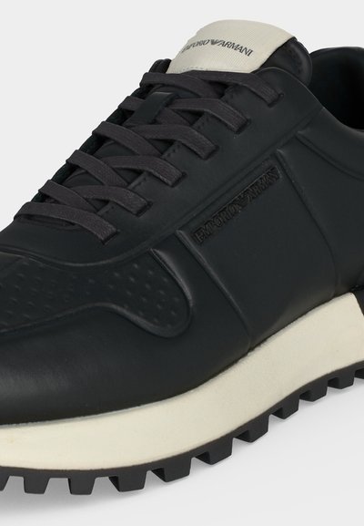 Baskets noires Emporio Armani avec lacets noirs, semelle blanche, logo de la marque en relief sur le côté et logo blanc sur la languette.