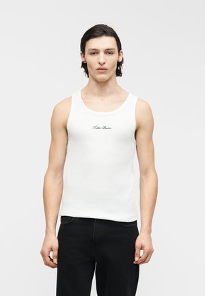 Young man with dark hair wearing a white sleeveless shirt with text and black pants, standing against a plain light background.