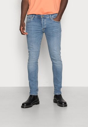 Homme portant un jean slim bleu clair, des bottes en cuir noir et une chemise orange, debout les mains dans les poches.