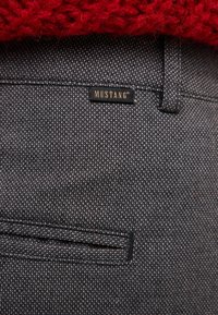 Grey textured fabric pants with a visible pocket and a black 'MUSTANG' tag. Above, a red knitted garment is partially visible.