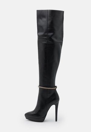 Botte noire en cuir montante jusqu’au genou avec talon stiletto, semelle plateforme et chaîne dorée décorative autour de la cheville.
