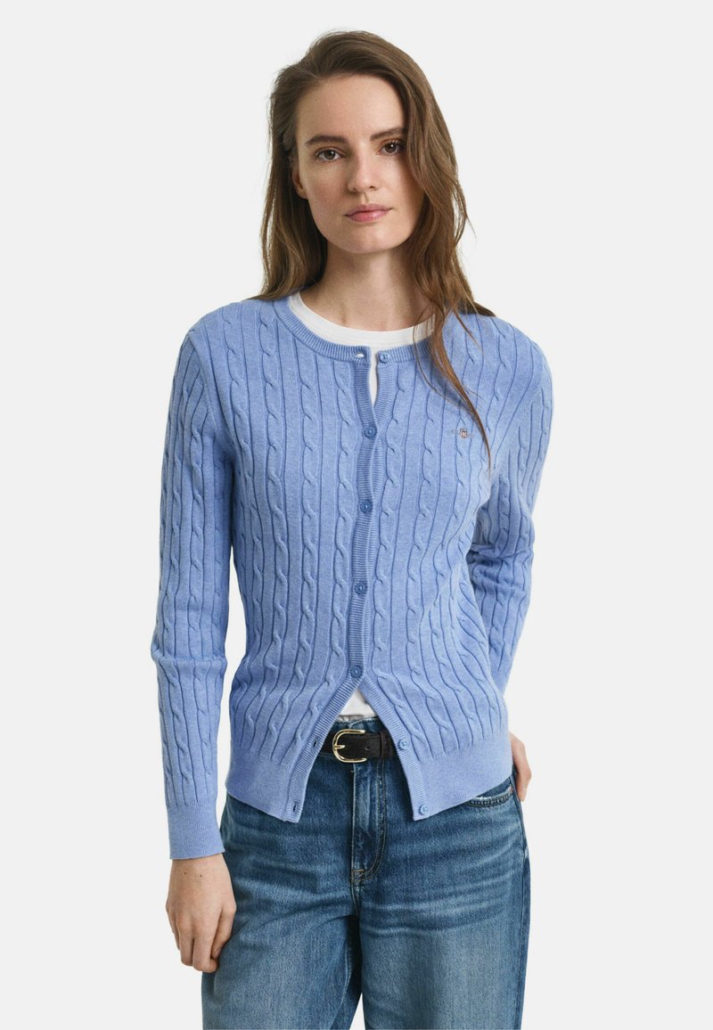 Svetlomodrý pletený kardigan s dlhými rukávmi a zapínaním na gombík. Má rebrovaný golier a lem. Nosený cez biele tričko a tmavé dž�ínsy.