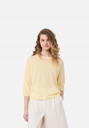 Giovane donna con capelli ricci indossa un top lavorato a maglia giallo chiaro con maniche 3/4 e pantaloni bianchi larghi, in piedi con una mano in tasca.