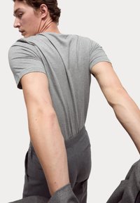 Šedé tričko s krátkým rukávem, bavlněný materiál, přiléhavý design. Model nosí šedé kalhoty na míru, drží šedý blazer, ukazující moderní vzhled.
