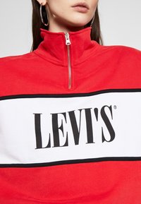 Rood sweatshirt met hoge kraag en een witte logoblok waarin "LEVI'S®" in vette zwarte letters staat. Voorzien van een zilveren rits bij de hals.
