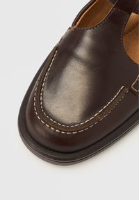 Chaussure en cuir marron avec un bout arrondi, présentant des coutures blanches contrastantes et un design ouvert. Texture lisse avec des embellissements minimaux.