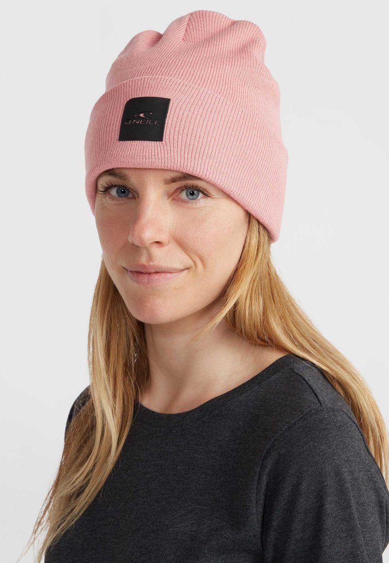 Beanie rosa a coste con un'etichetta logo rettangolare nera. Il materiale appare morbido e la vestibilità è aderente con una sezione superiore leggermente sovradimensionata.