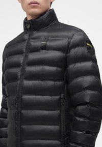 Homme portant une doudoune noire avec col montant, fermeture éclair devant, poches latérales zippées et logo jaune Blauer sur la poitrine et sur le patch de la manche.
