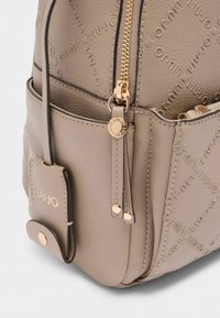 Zaino in pelle texturizzata beige con logo impresso, hardware dorato, tasca laterale e chiusura con zip. Presenta un dettaglio pendant.