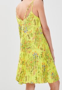 Une robe florale jaune avec des bretelles fines, dotée de plis et d'un dos en V. Le tissu présente des motifs vibrants de feuilles et de fleurs dans diverses couleurs.