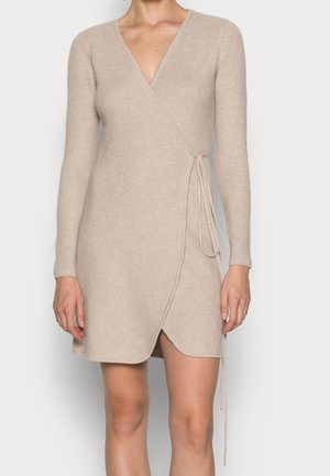 Femme porte une robe portefeuille en maille côtelée beige avec manches longues et nœud sur le côté, terminant au-dessus du genou.