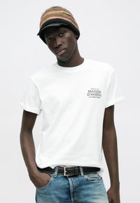 Noor mees kandmas triibulist kootud mütsi ja valget Maison D'Hiver t-särki, mis on pistetud sinistesse teksapükstesse musta vööga, poosistades ühe käega püksitaskus.