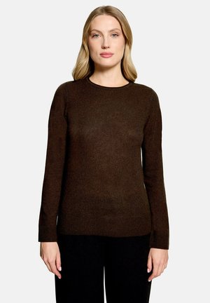 Femme aux cheveux blonds portant un pull marron foncé à manches longues et un pantalon noir, debout devant un fond clair uni.
