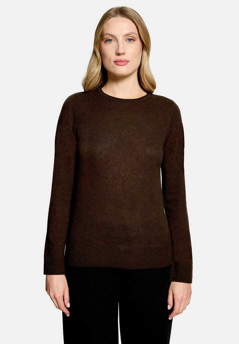 Femme aux cheveux blonds portant un pull marron foncé à manches longues et un pantalon noir, debout devant un fond clair uni.