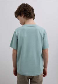 T-shirt en coton turquoise à manches courtes, col rond et texture lisse. La vue arrière montre une coupe décontractée et une petite étiquette à l'ourlet.