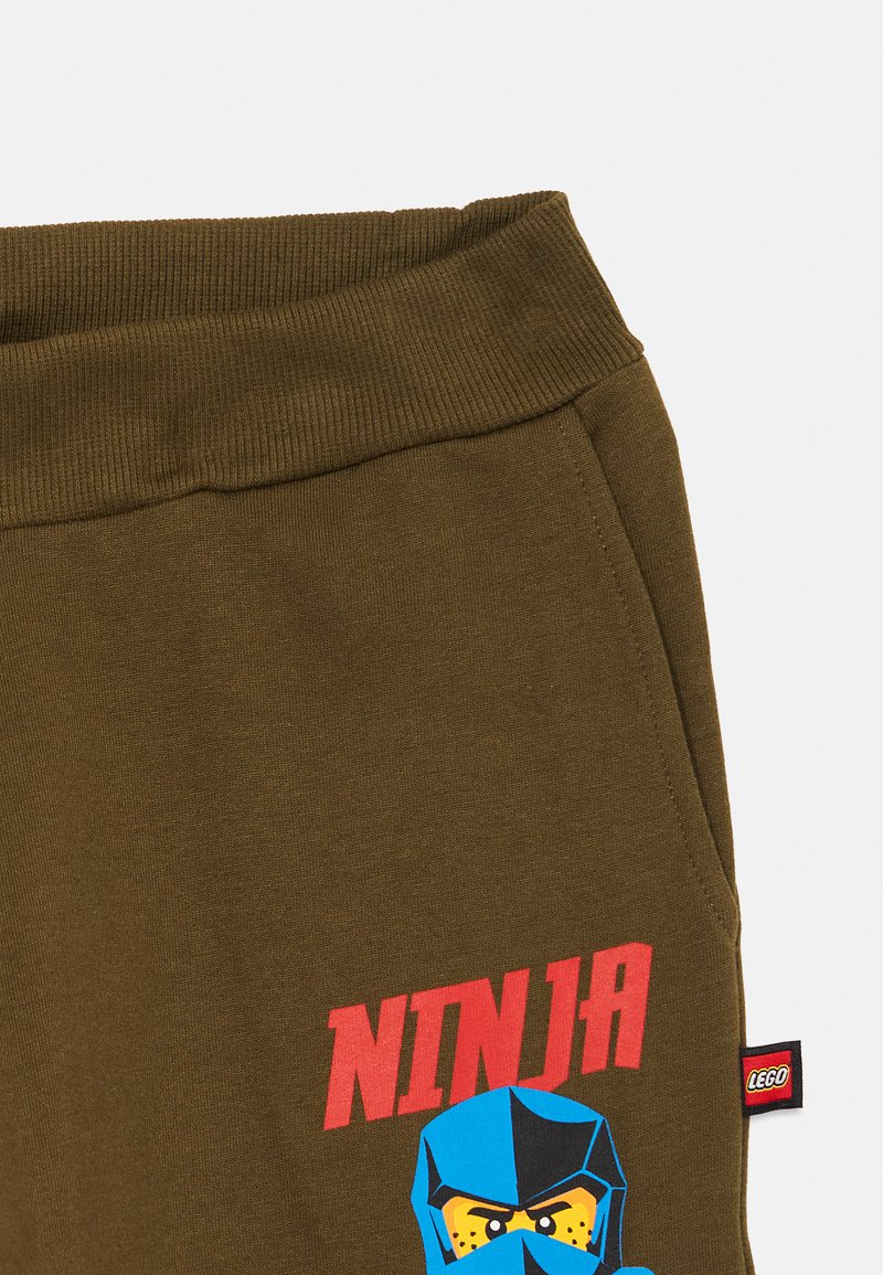 Olivgrüne Sweatpants mit geripptem Bund, mit einem blauen Ninja-Graphic und dem Wort "NINJA" in Rot. Mit einem LEGO-Tag.
