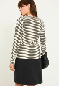 Langärmliges Oberteil mit schwarz-weißen horizontalen Streifen; kombiniert mit einem dunklen, strukturierten Rock; das Model hält eine schwarze Handtasche.