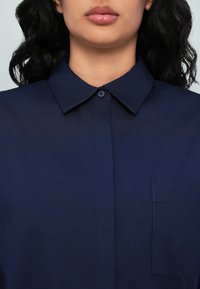 Femme au teint moyen et aux cheveux noirs ondulés portant une chemise bleu marine boutonnée avec un col et une poche poitrine.