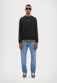 Svart genser med grønn innvendig lag, crew neckline og hvite sidestreker. Bæres med lyseblå jeans og grå sneakers.