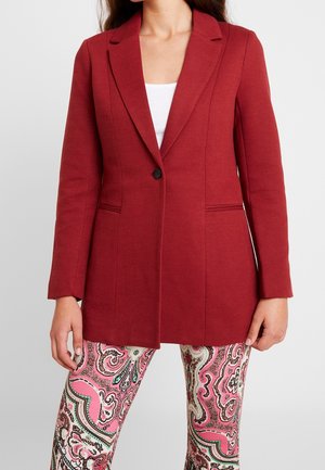 Kobieta ubrana w długi, czerwony jednorozporkowy blezer na jeden guzik, na biały top, w połączeniu z różowymi spodniami w wzór paisley.