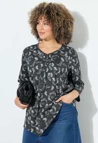 Mujer con cabello rizado, vestida con una camisa de manga larga negra con estampado y una falda de mezclilla azul, sosteniendo un bolso de mano negro, de pie contra una pared blanca.