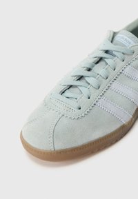 Világoskék szarvasbőr Adidas tornacipő három fehér csíkkal, hozzáillő fűzőkkel és egy gumitalpú talppal, fehér háttér előtt.