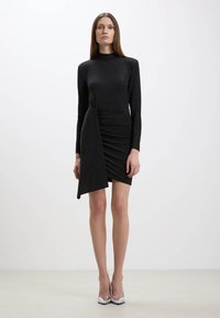 Abito lungo nero a maniche lunghe con collo alto, caratterizzato da un davanti drappeggiato e tessuto strutturato, abbinato a tacchi alti metallici.