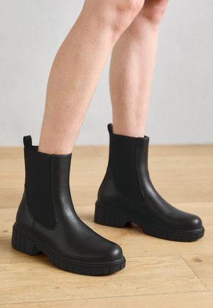 Černé kotníkové boty vyrobené z hladkého matného materiálu, s elastickými bočními panely, chunky podešví a poutkem na zadní straně pro snadné obouvání.