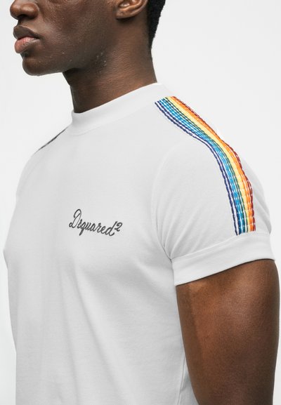 Κοντινό πλάνο ατόμου που φοράει λευκό πουκάμισο με κοντό μανίκι και πολύχρωμες ρίγες στον ώμο, με το λογότυπο "Dsquared²" στο στήθος.