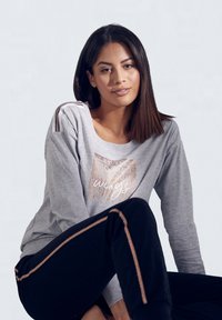 Ung kvinde med glat mørkt hår iført en grå sweatshirt med metallisk tryk og sorte bukser med kobberfarvede sidestriber, siddende mod en ensfarvet baggrund.